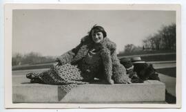 Woman reclining on concrete, wearing a geometric patterned skirt and bulky shearling coat, with trees in background.
