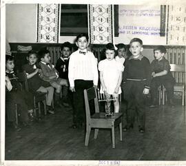 Workmen's Circle Abraham Reisen kindergarten. Interior group portrait of two male students and a female student standing behind a chair with a lit menorah. Other students seated in background.